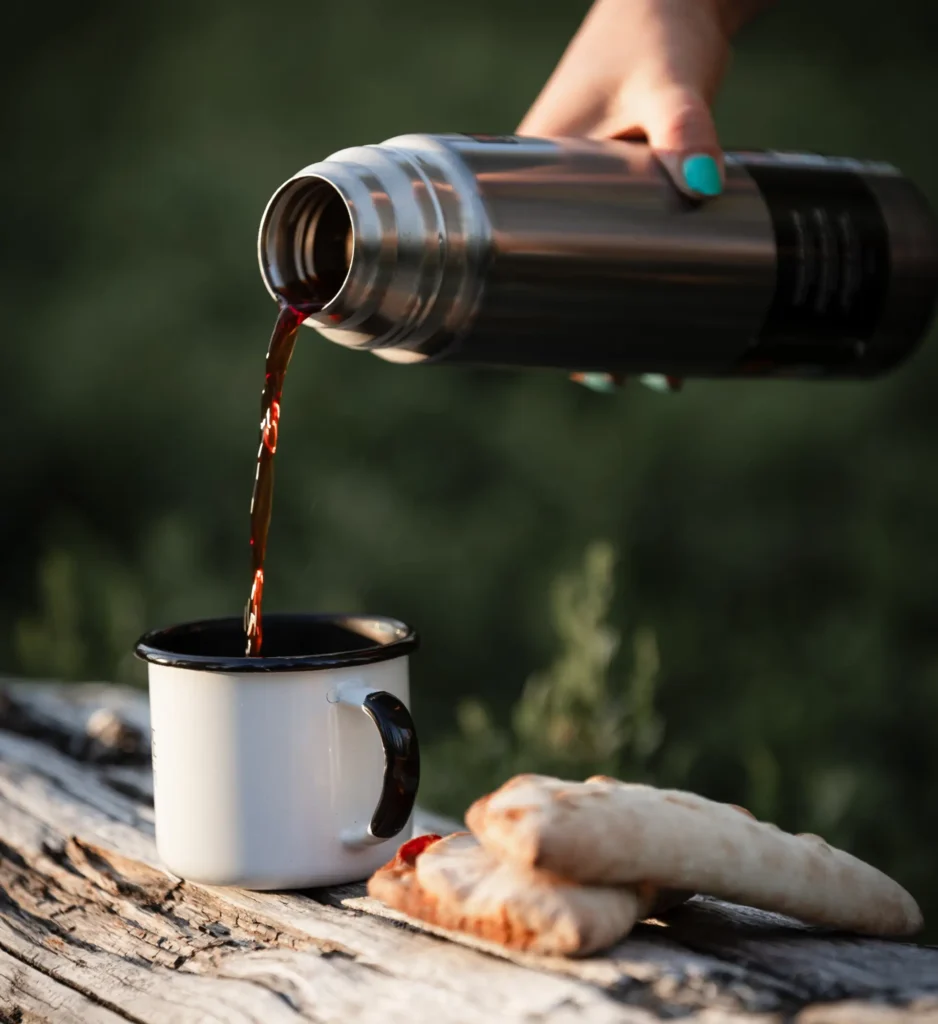 Coffee being poured from a thermos into an enamel mug with Finnish pastries outdoors.