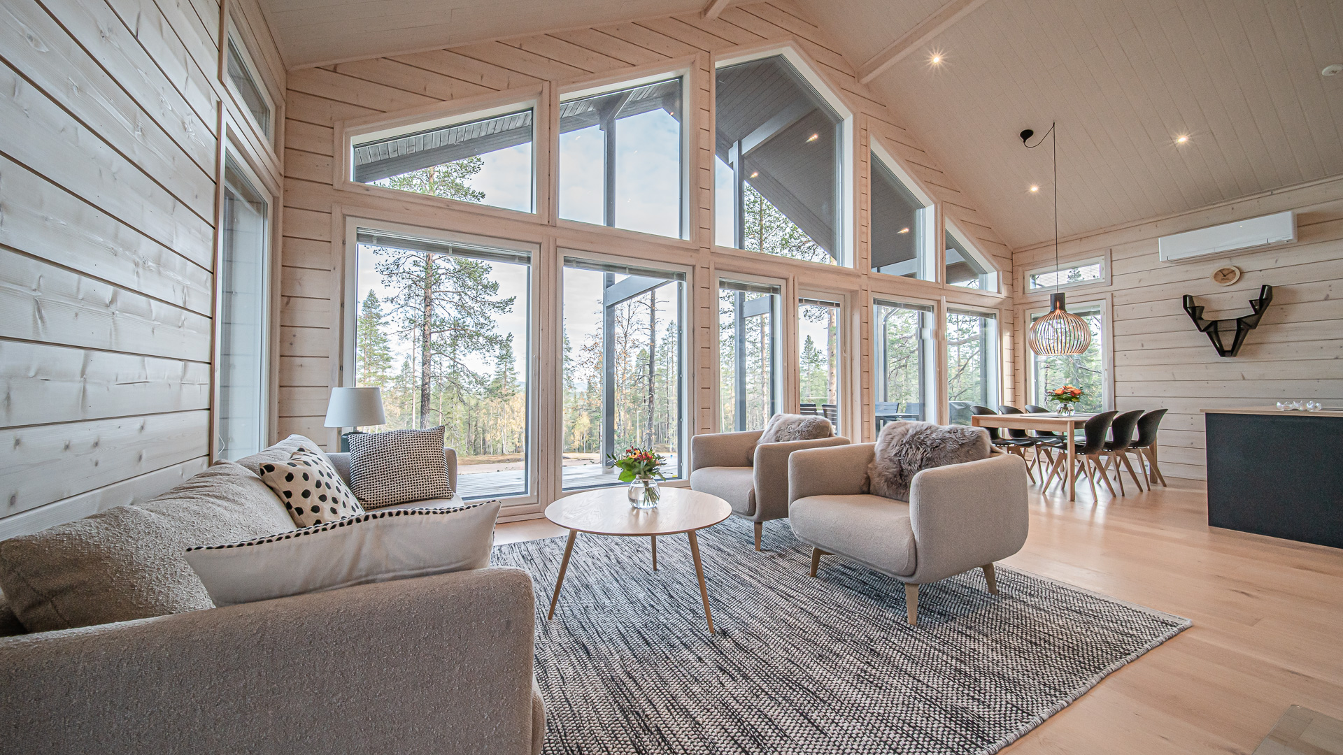 A cabin with large windows showcasing a forest landscape. Grey sofa and armchairs, and a dining table for 6 people.