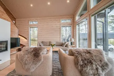 Moderni ja valoisa olohuone, jossa on suuret ikkunat, vaaleat hirsiseinät, takka, beige sohva ja kaksi nojatuolia taljoilla.