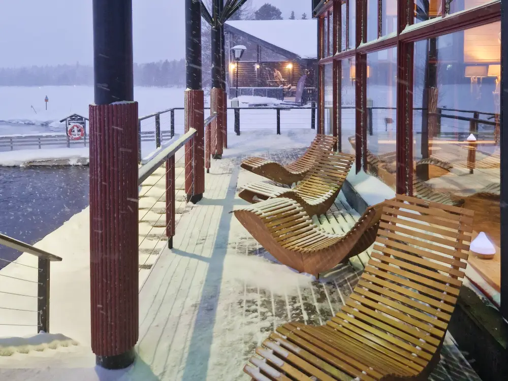 arcticsaunaworld Snow-covered wooden lounge chairs on the terrace of Arctic Sauna World in Muonio, overlooking a frozen lake in a peaceful winter setting.