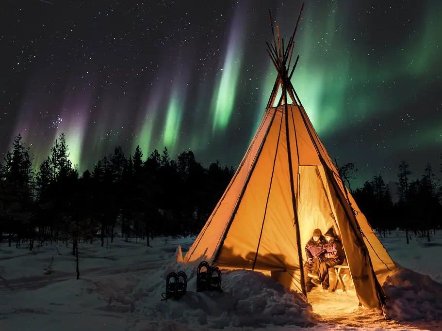 northern_lights Two people sit inside a warmly lit traditional tipi tent, Finnish kota, in a snowy forest at night, with vibrant green and purple northern lights illuminating the starry sky above.