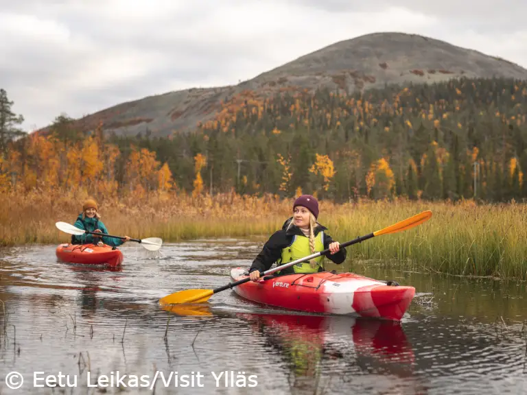 Yllas ruska melonta kesankijoki EetuLeikas-VisitYllas Two people kayaking on a calm river surrounded by autumn-colored trees in Ylläs, Finland, with a fell rising in the background.