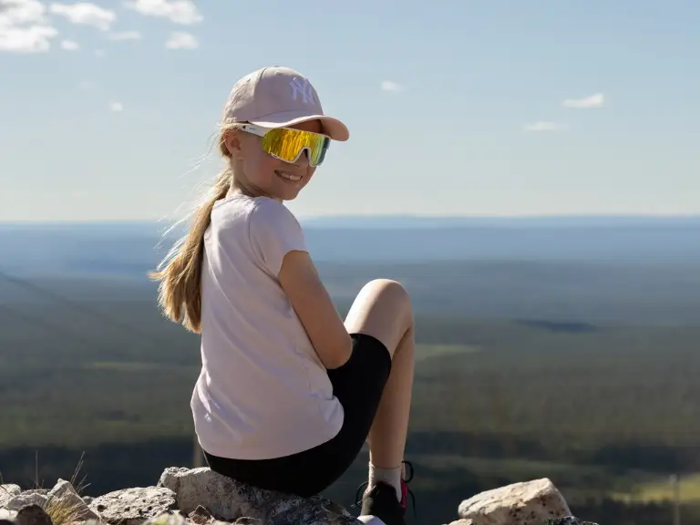 yllas_huippu A girl sitting on top of Ylläs, wearing a cap and sunglasses, smiling at the camera.