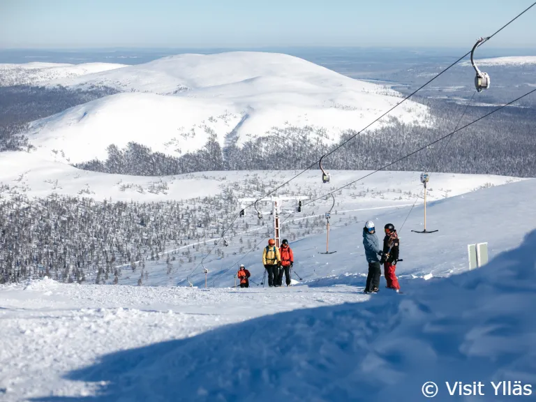 Yllas hankikanto laskettelu YllasSkiResortNorth c Skiers using a T-bar lift on the snowy slopes of Ylläs Ski Resort, with wide arctic fells and forested valleys in the background under a clear blue sky.