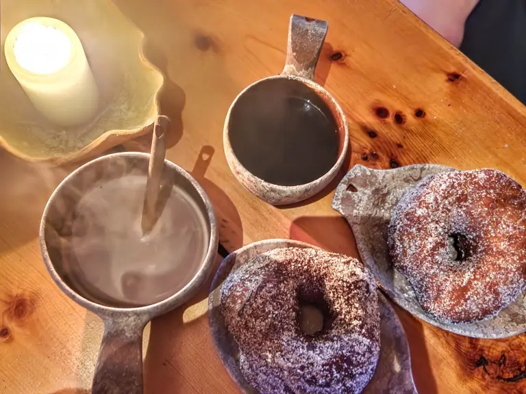 yllas cafes Traditional wooden cups filled with steaming hot chocolate and coffee, served with sugar-coated doughnuts on a wooden table next to a lit candle.