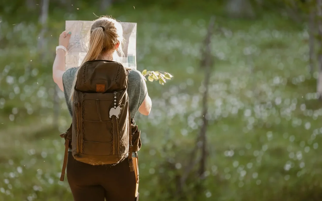 A person with a backpack stands in a sunlit forest, facing away from the camera and holding a map while navigating outdoors.