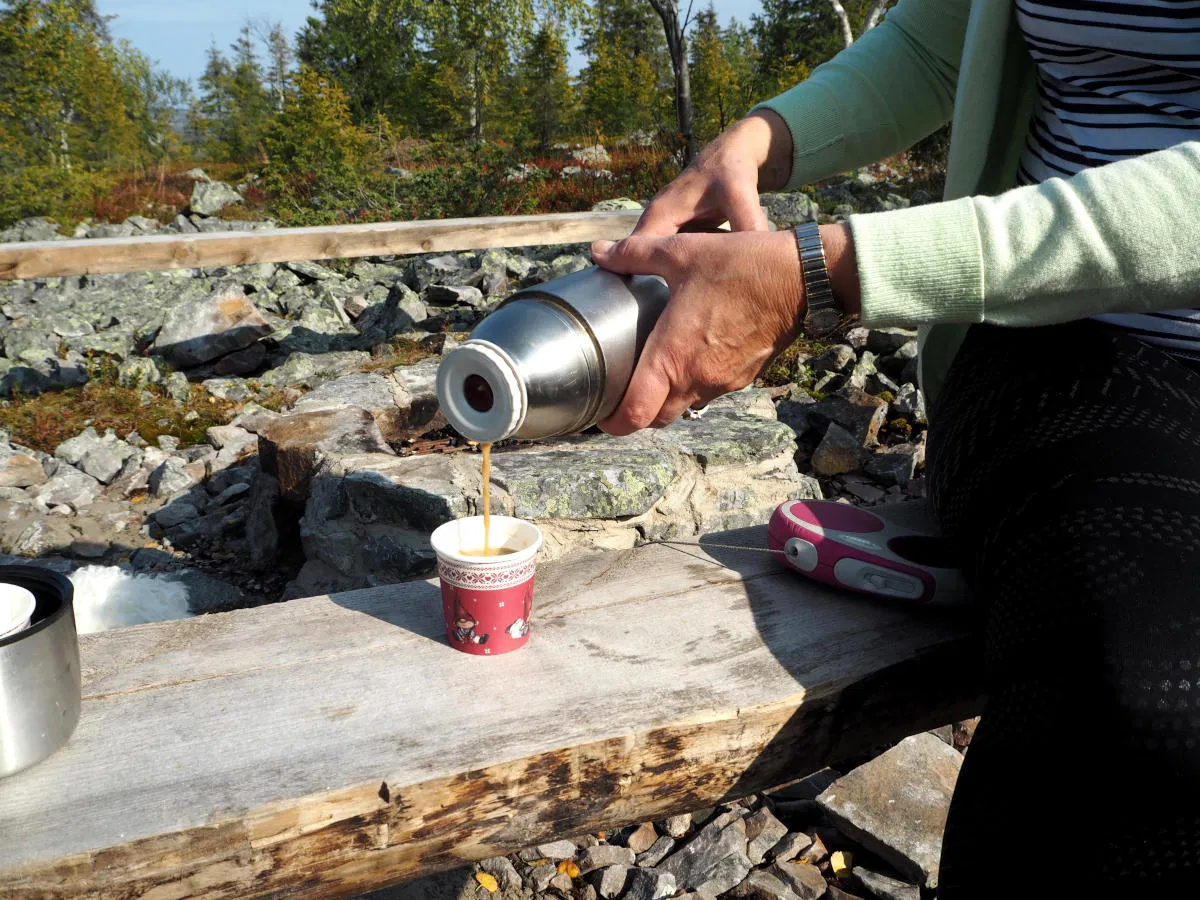 Person pouring coffee from a thermos into a paper cup at a scenic rest stop in Finnish Lapland.